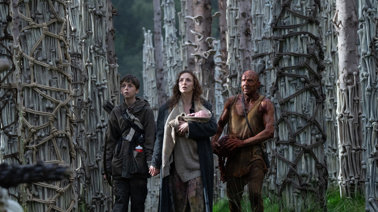 Spike, Isla (holding a baby), and Dr. Ian Kelson standing among many bones in 28 Years Laterr